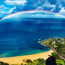 A rainbow arches over a beach with turquoise waters and green hills under a partly cloudy sky.