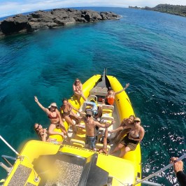 People relaxing on a yellow boat in clear blue water near rocky coast.
