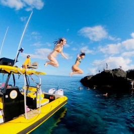 Two people jump off a yellow boat into clear blue water.