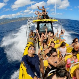 Group of people smiling and waving on a yellow boat in the ocean.
