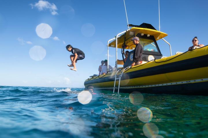 Person jumps off yellow boat into the ocean, with others watching under a clear blue sky.