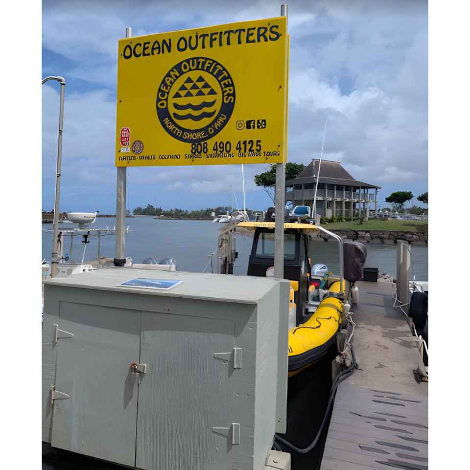 Docked boat with 'Ocean Outfitters' sign and a yellow raft, water and house in the background.